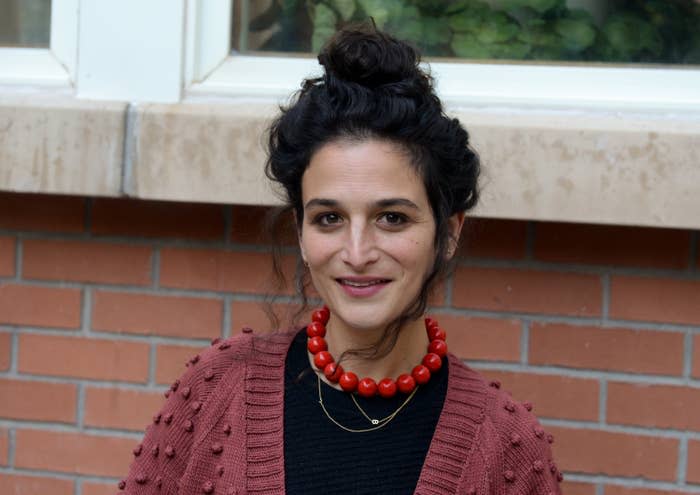 Jenny Slate smiles while posing for a photo outside and in front of a brick wall