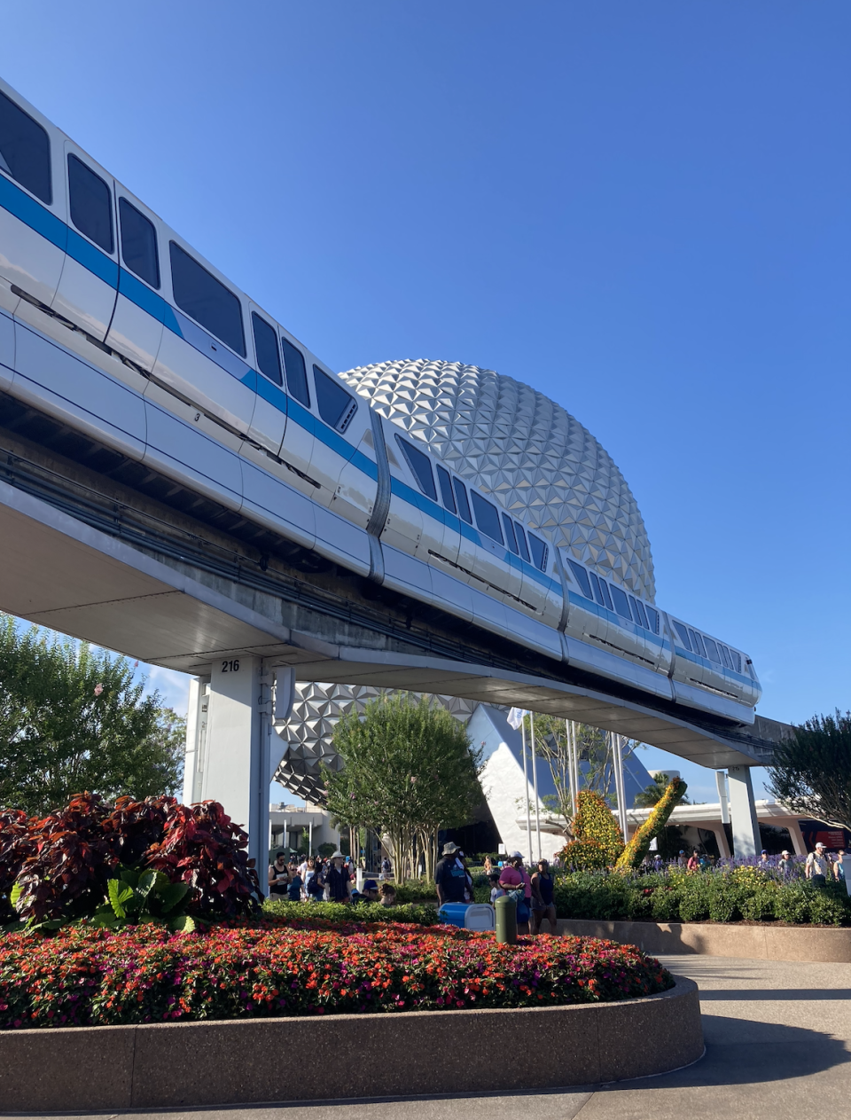 Monorail passes by Epcot's Spaceship Earth with vibrant flowers and visitors in the foreground
