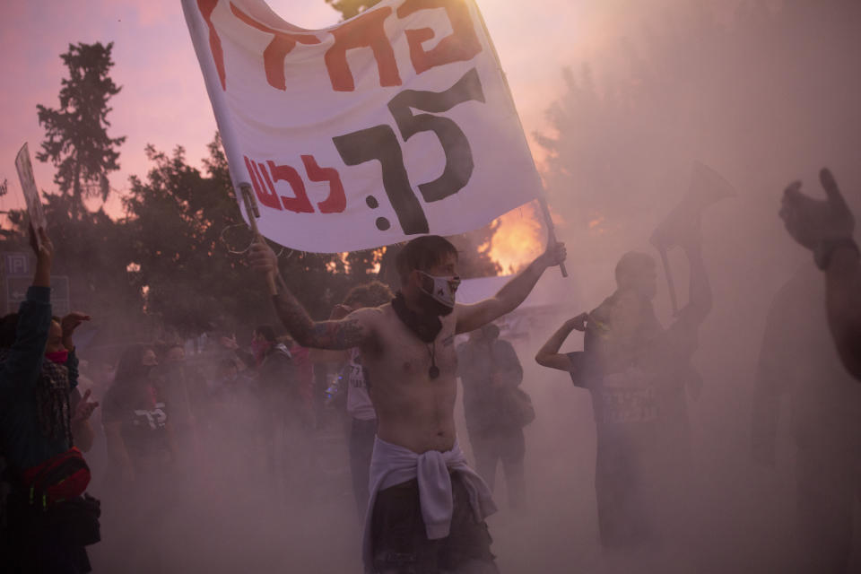 An Israeli anti-government protester holds a banner as he is enveloped in the smoke from a bonfire outside of the official residence of Prime Minister Benjamin Netanyahu on the day his corruption trial was originally scheduled before it was postponed, in Jerusalem, Wednesday, Jan. 13, 2021. In Hebrew, the sign reads: "Coward! Go to trial!" (AP Photo/Maya Alleruzzo)