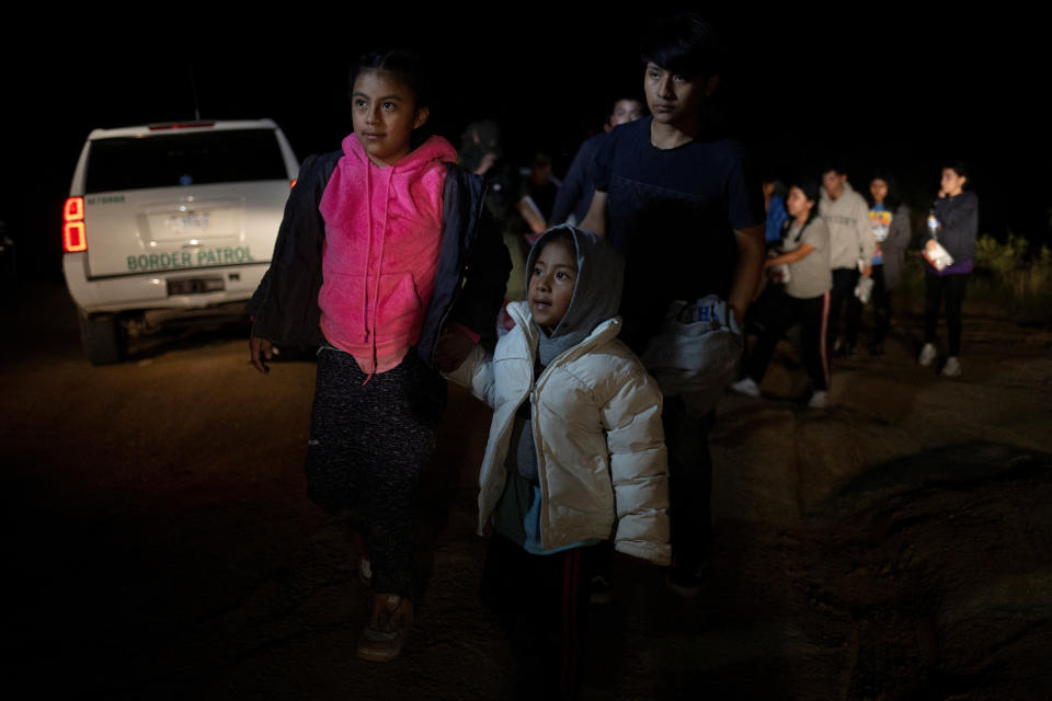 Two migrants from Guatemala traveling without adults, one of about 5 and the other about 11, walk toward a border patrol transport vehicle, with a young man walking behind them.