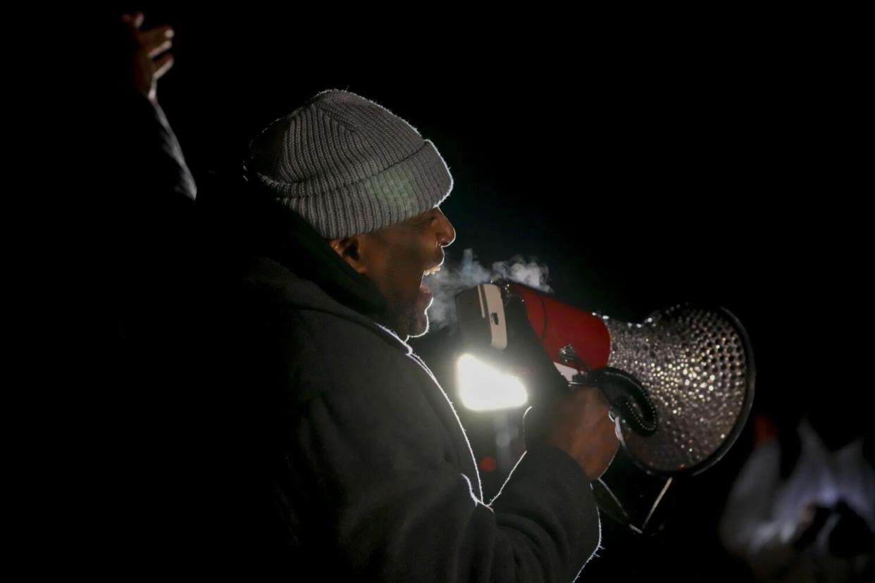 A man speaking into a megaphone in the dark, his breath visible in the cold