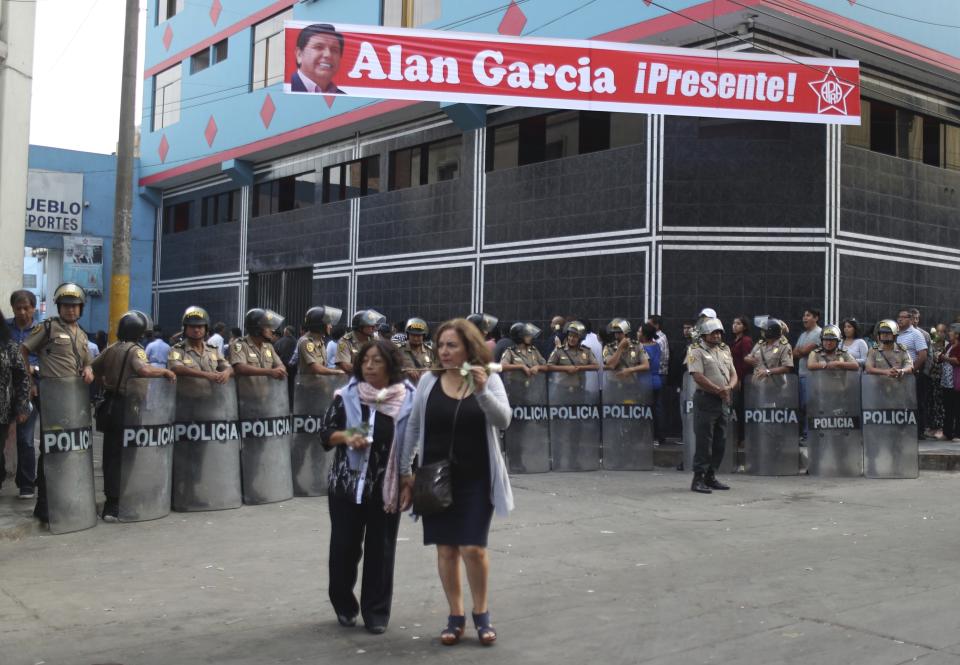 A sign featuring Peru's late President Alan Garcia reads in Spanish "Alan Garcia is present!" as police stand guard near his political party's headquarters where women carrying roses walk to his wake there in Lima, Peru, Friday, April 19, 2019. Garcia shot himself in the head and died Wednesday as officers waited to arrest him in a massive graft probe that has put the country's most prominent politicians behind bars and provoked a reckoning over corruption. (AP Photo/Martin Mejia)