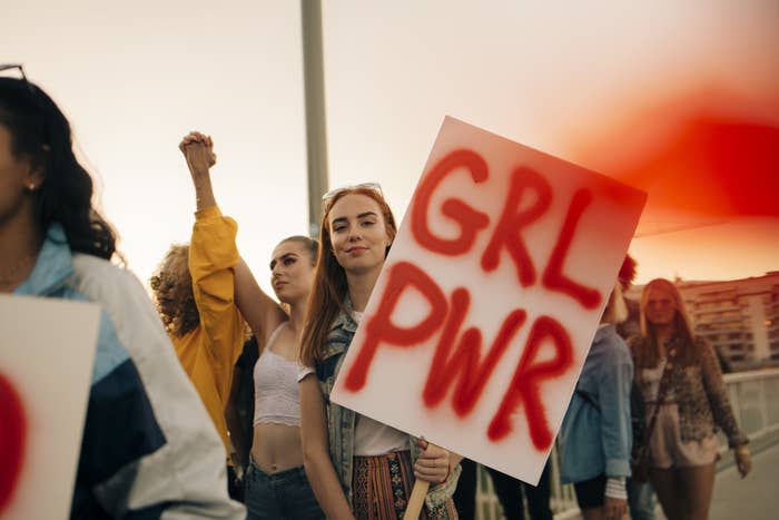 Group of people at a protest, one holding a "GRL PWR" sign. In the foreground, one person has their fist raised