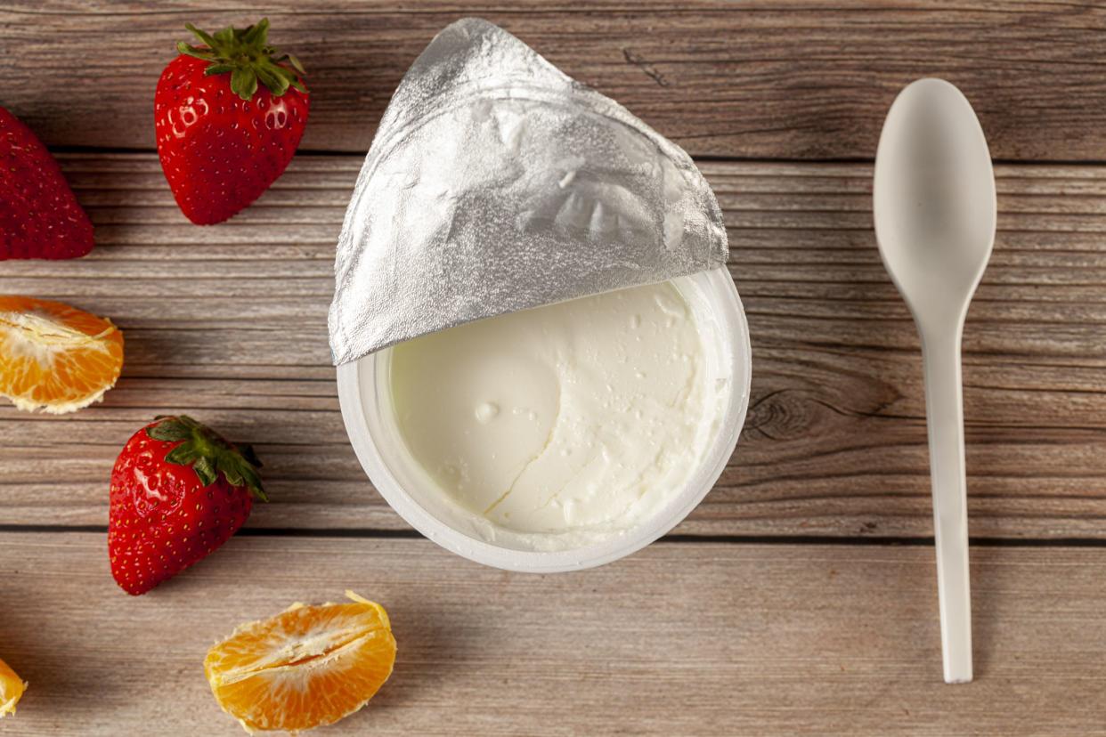 Flat lay image of a store bought plastic cup of plain low fat Greek Yogurt with the aluminum foil seal peeled off. It is on wooden background with a plastic spoon and fresh fruits. A diet idea concept