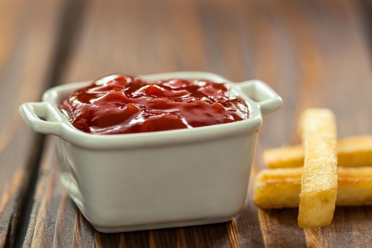 Closeup of ketchup in a white dipping bowl with a few french fries on a wooden table with a blurred background