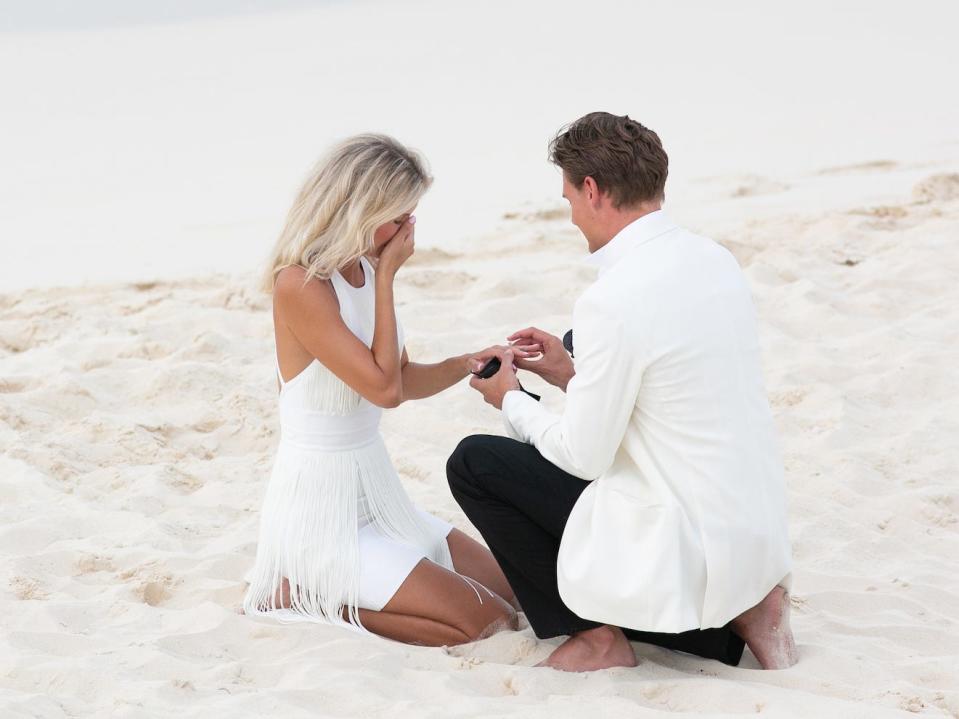 A man in a white jacket on his knees puts a ring on a woman's finger as she kneels next to him.