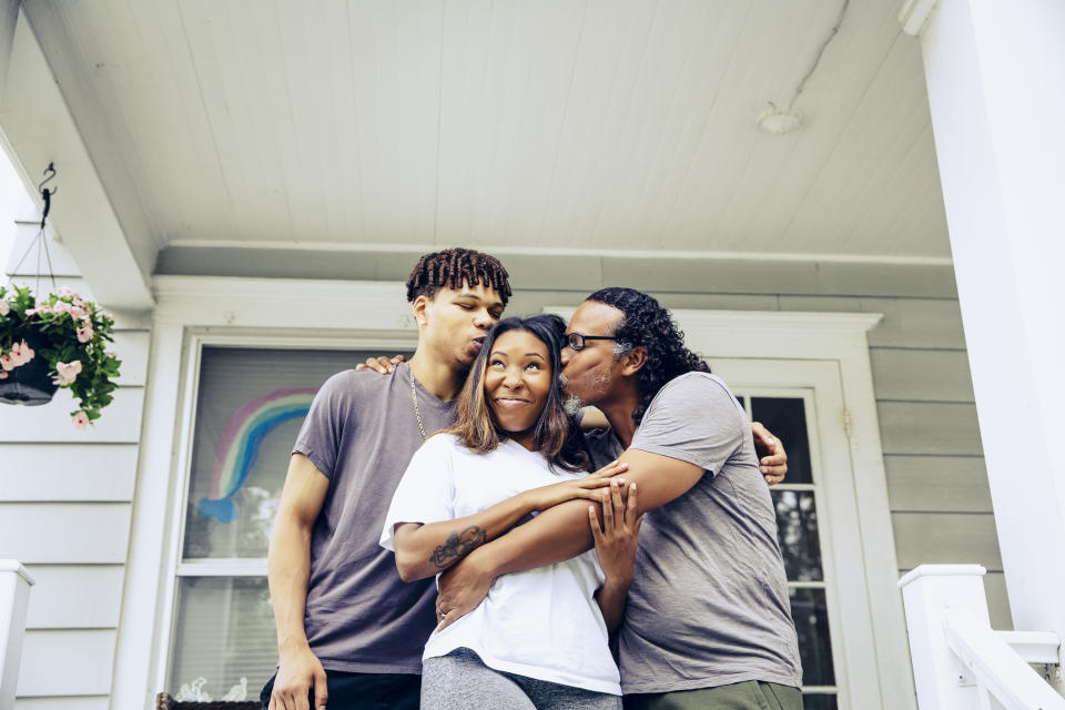 A family of three, with a man and a young man kissing a woman on either cheek, stands on the porch of a house. A rainbow drawing is visible in the background