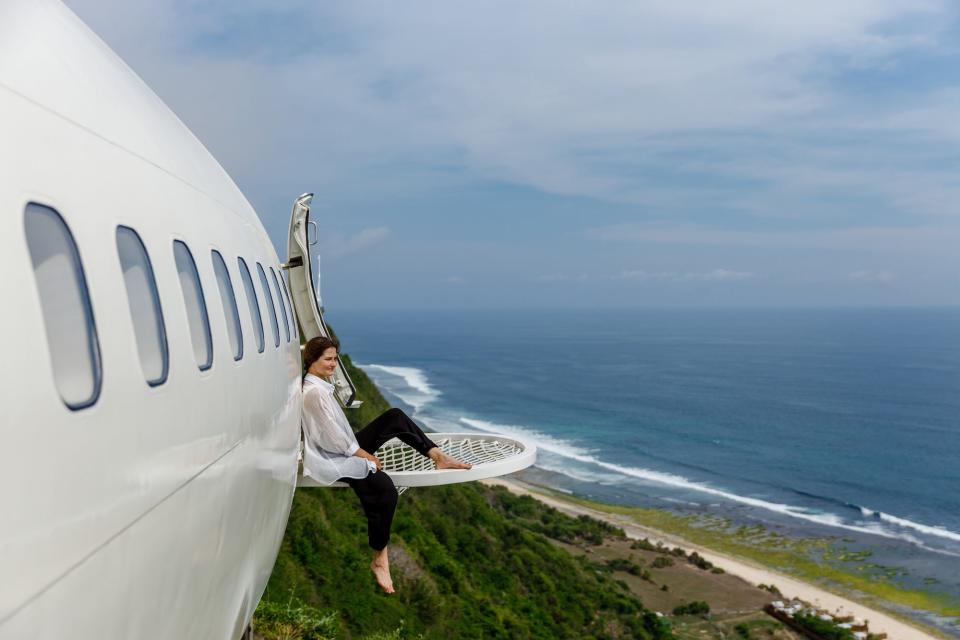 A woman sits on a hammock attached to the side of a white plane overlooking a beach.