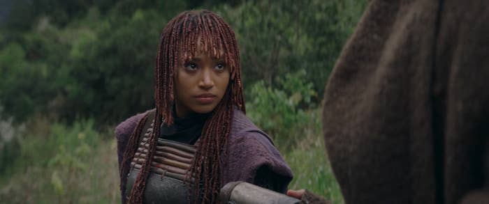 Amandla Stenberg dressed in a detailed, futuristic outfit, facing another person in an outdoor, forest-like setting