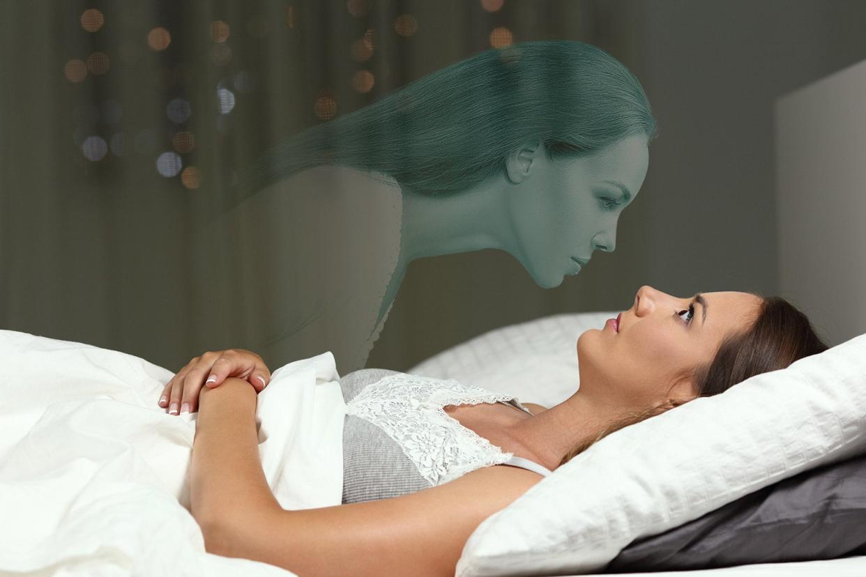 A woman lying in bed stares wide-eyed at an apparition of a woman hovering over her. 