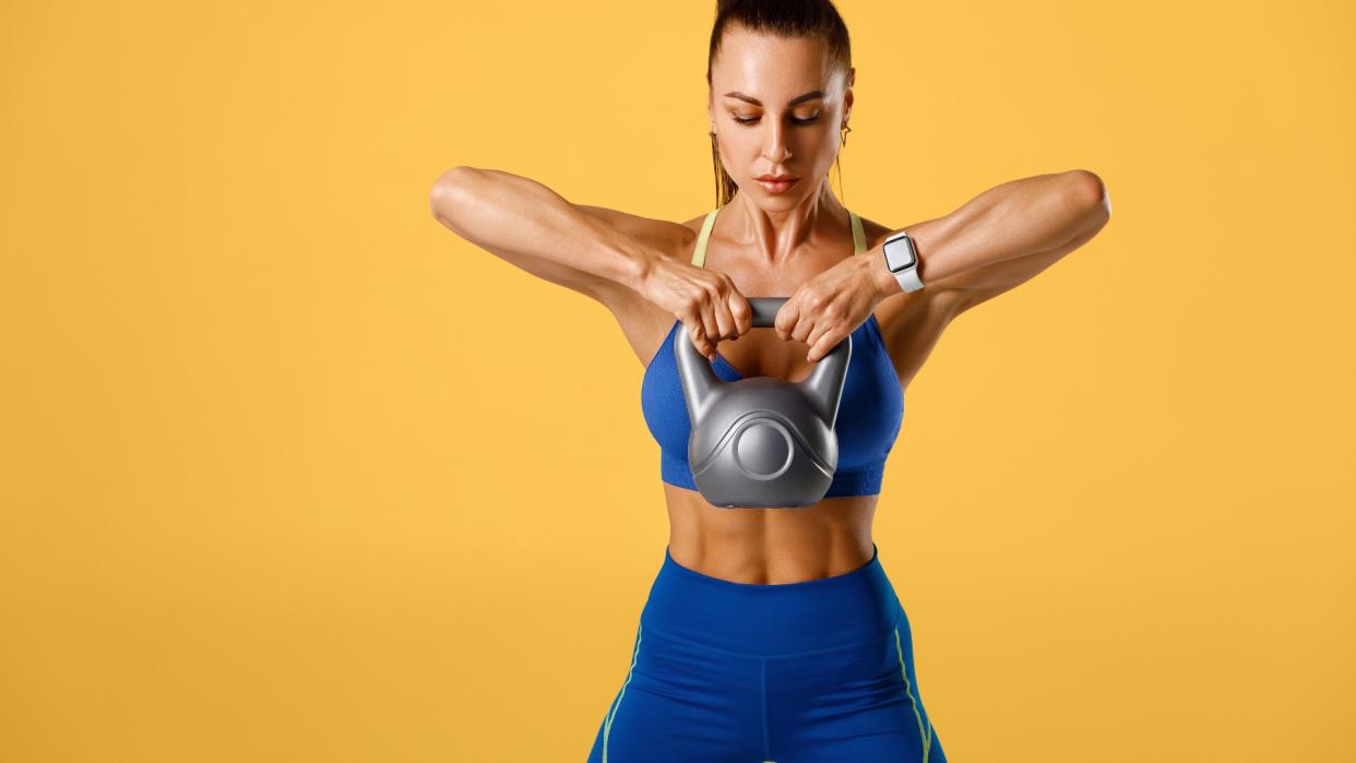  Woman performing an upright row holding a kettlebell up to her chest against a white background, showing abs 