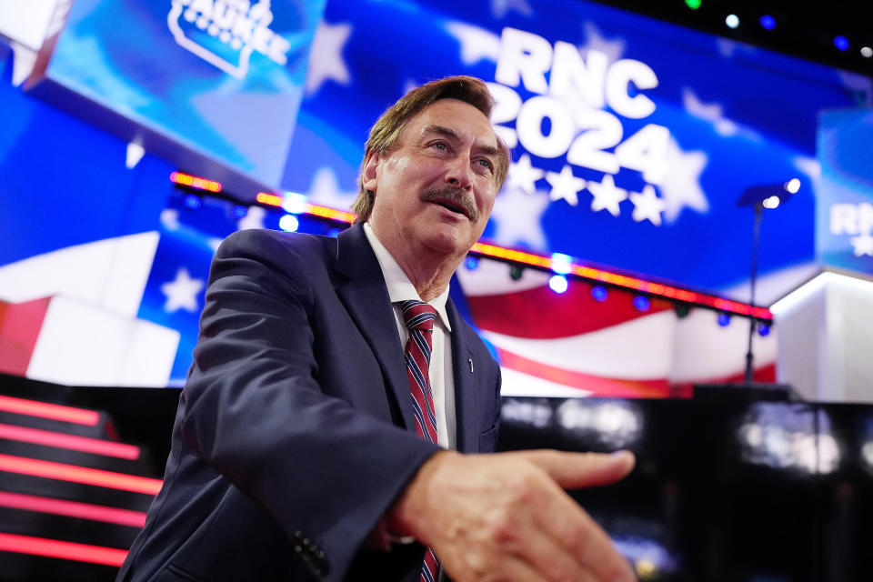 Mike Lindell streckt seine Hand auf der Bühne der RNC 2024-Veranstaltung aus. Er trägt Anzug und Krawatte. Im Hintergrund sind Elemente der amerikanischen Flagge zu sehen und "RNC 2024" Text