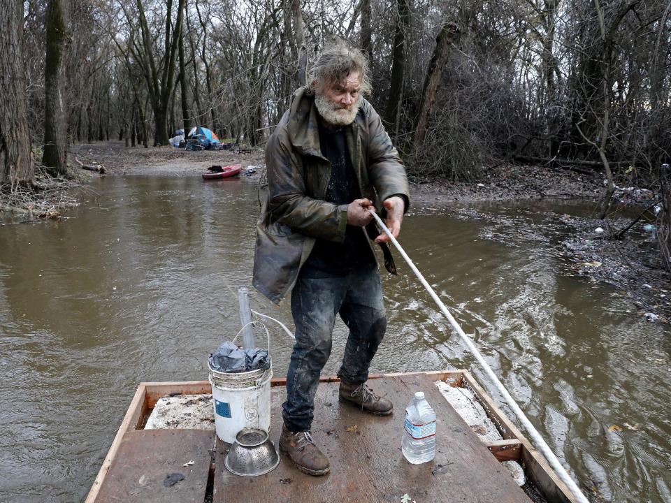 bearded man wearing rain jacket pulls rope on wooden raft in flooded forest river with tent in background
