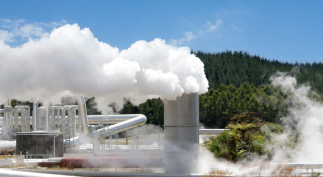 A geothermal power plant operates with a forest and a clear, bright sky visible behind it.