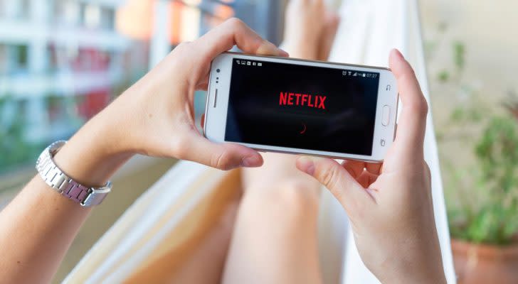 Picture of a person laying on a couch holding a mobile phone that features the Netflix (NFLX) logo on the screen