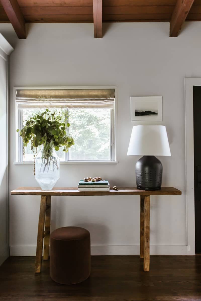 red wood beams on ceiling, simple wood entry table, beige roamn style curtains, large base table lamp with white shade