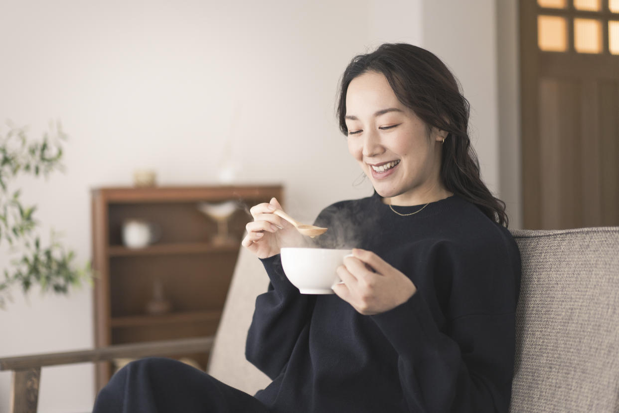 An Asian woman is sitting on the sofa, holding a cup and drinking soup. She is wearing long sleeves.