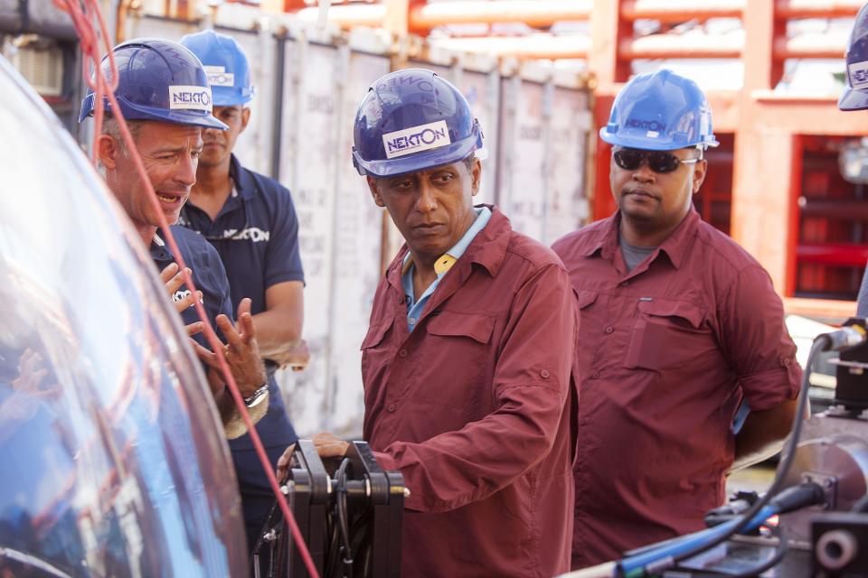 In this Saturday, April 13, 2019, photo, Seychelles President Danny Faure, center, gets briefed on the details of submersible operations on the deck of vessel Ocean Zephyr, off the coast of Desroches, in the outer islands of Seychelles. Faure toured the vessel and was presented with some of the findings and observations made by a British-led science expedition documenting changes taking place beneath the waves that could affect billions of people in the surrounding region over the coming decades. (AP Photo/Steve Barker)