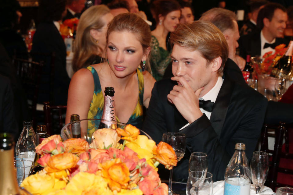 Taylor Swift in an elegant dress and Joe Alwyn in a tuxedo are seated at a formal event with a floral centerpiece