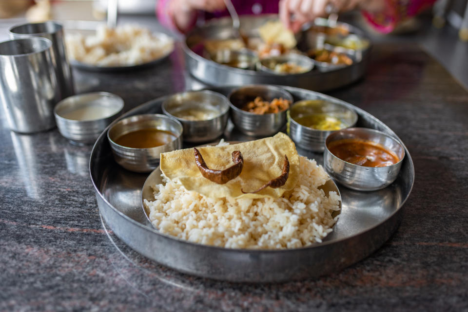 Indian thali with rice, various curries and a papadum on a metal tray, hands serving food in the background