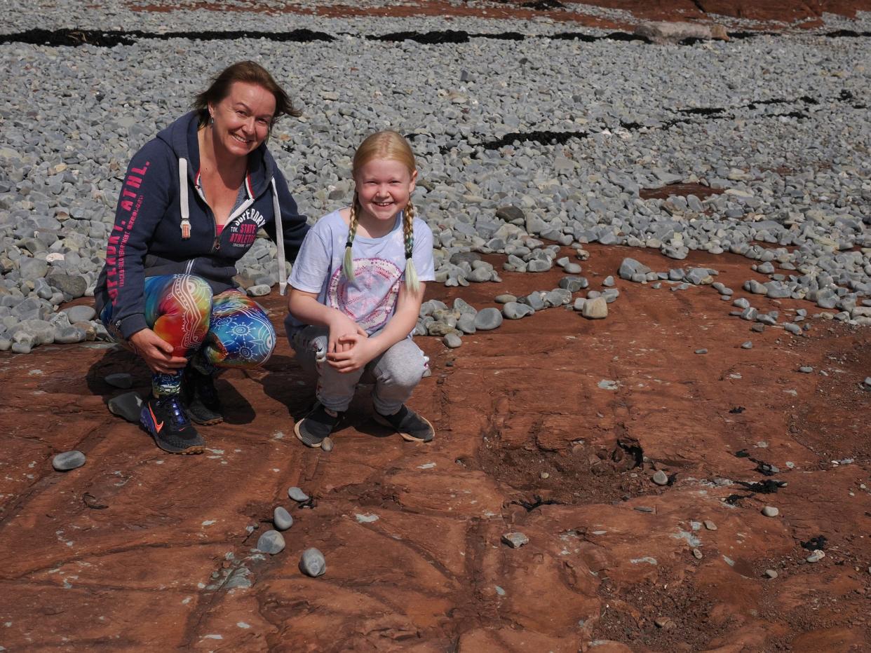 A woman and girl crouch and smile by dinosaur footprints on the beach