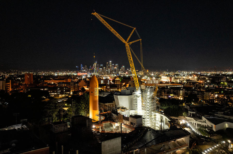 night view of a yellow crane lifting a large orange fuel tank oriented vertically.