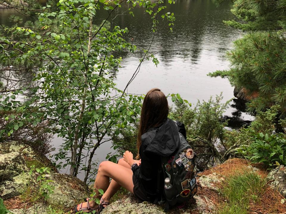 Emily, wearing a hooded jacket and a backpack covered in patches, sits on a grassy rock and looks out at the water and trees.