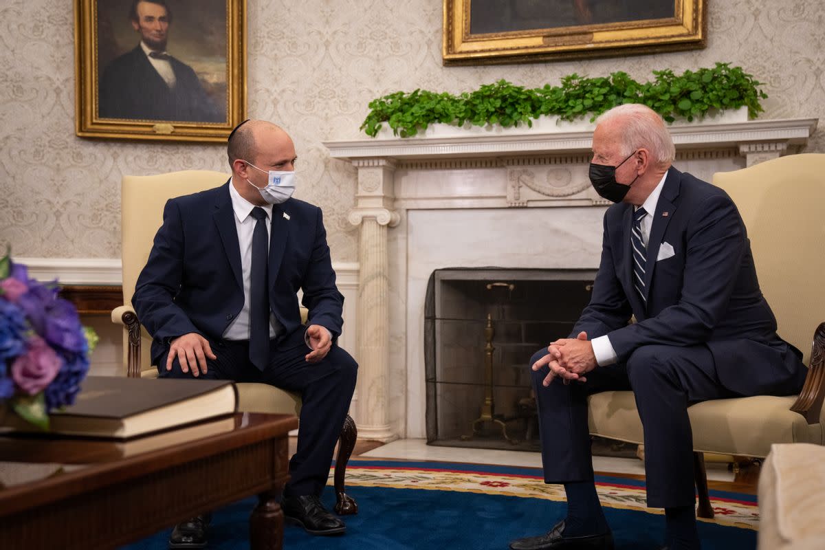 Two white men wearing masks and blue suits sit in white chairs. 