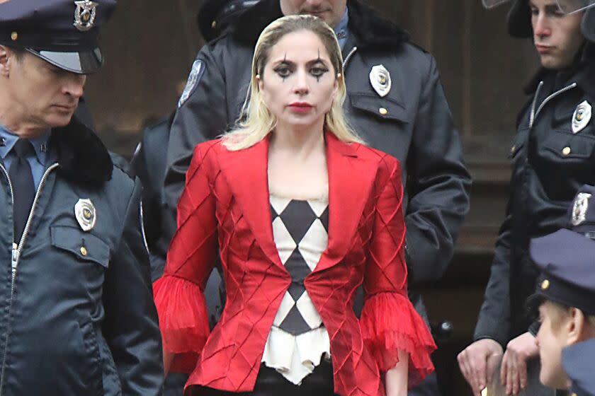 Lady Gaga wearing a red blazer, black-and-white blouse and clown makeup, surrounded by men in police uniforms