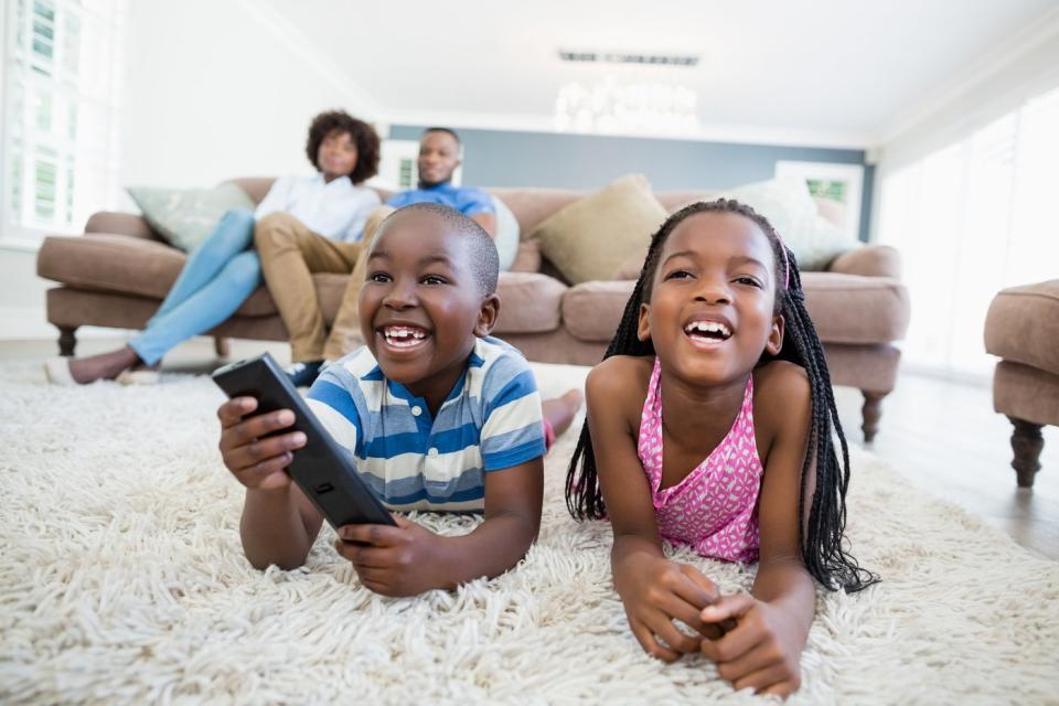 Two siblings are lying on a rug and watching TV, their parents are sitting on the sofa in the background.