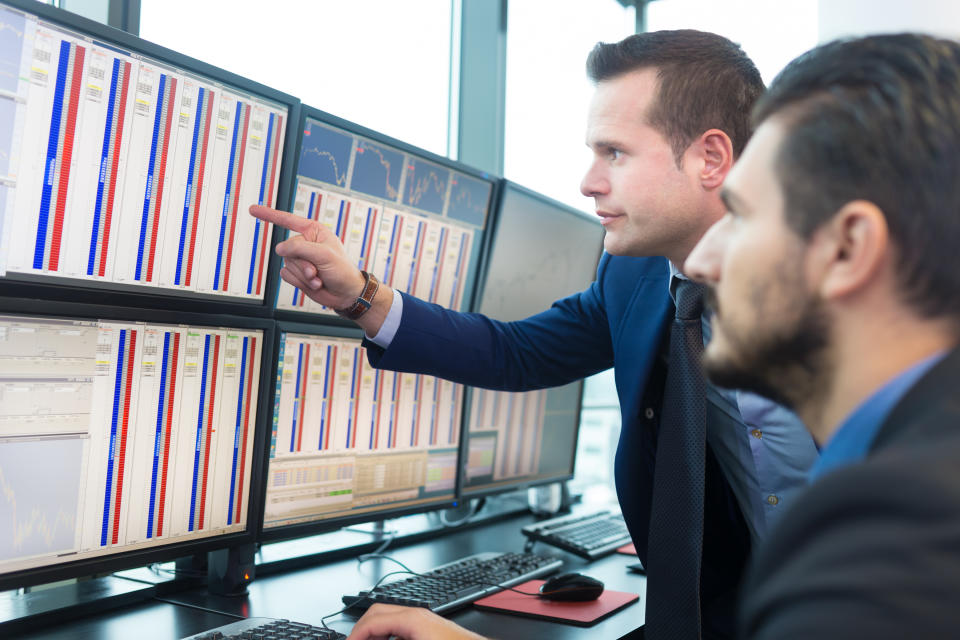 Two men in suits looking at charts on multiple computer screens.