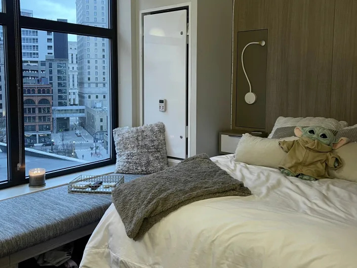 Bed with brown and white sheets and windowsill with view of Detroit apartment buildings