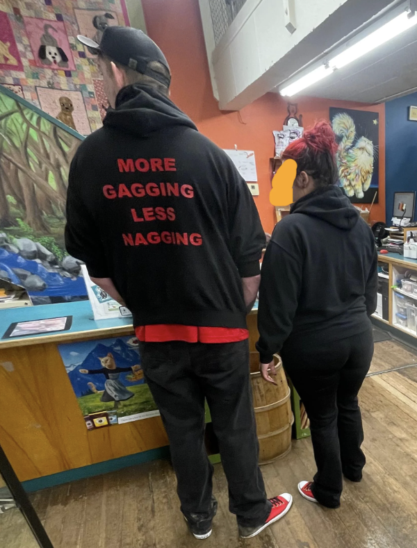 Two people standing in a shop; one has a sweatshirt with "MORE GAGGING LESS NAGGING" text on the back