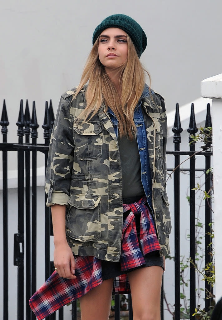 Cara Delevingne is seen in a casual outfit wearing a green beanie, camo jacket over a denim shirt, green t-shirt, and plaid shirt tied around her waist