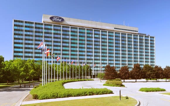 Ford's headquarters, a 12-story blue-glass-faced office building, viewed from a flag-lined driveway in front.