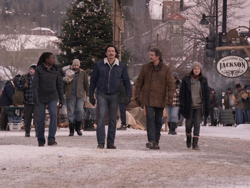 maria, tommy, joel, and allie walking on a snow covered road in the last of us, wiht a sign to the right that reads welcome to jackson