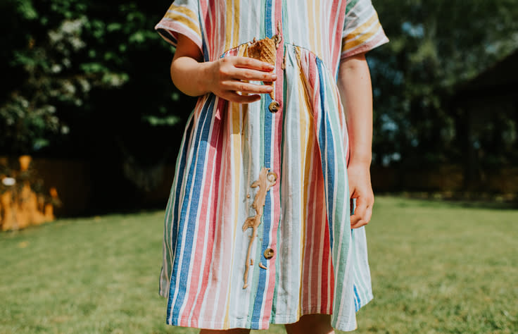 A young person standing on grass, wearing a striped dress with a large stain on the front. The person's face is not visible