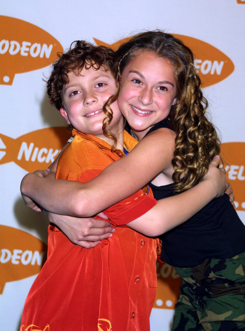 Spy Kids Daryl Sabara, in a red and orange shirt, and Alexa Vega in a dark singlet and green camo pants, hug on the red carpet with the Nickelodeon logo on the wall behind.