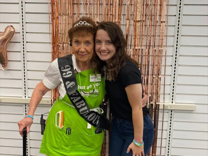 Jayne Burn, wearing a green JoAnn's apron, poses with her co-worker, Maggie HarsVar, in front of a glitter decoration during her 99th birthday party.