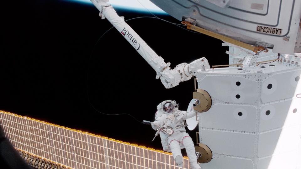 an astronaut beside a space station module and above a solar panel during a spacewalk. above him is a robotic arm with the word 