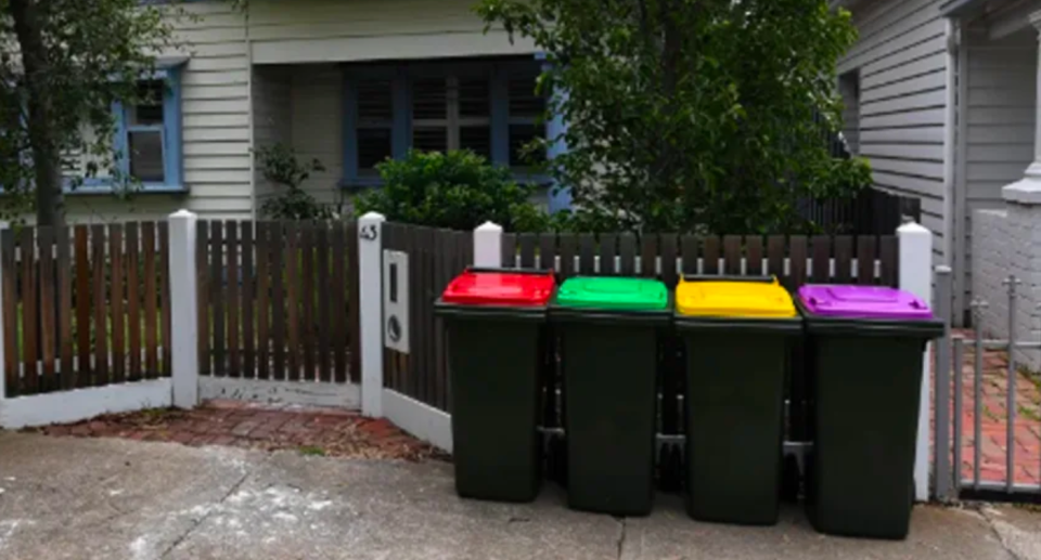 Photo of four bins lined up in front of a home. Bins, in order from left to right, have a red lid, green lid, yellow lid, and purple lid. 