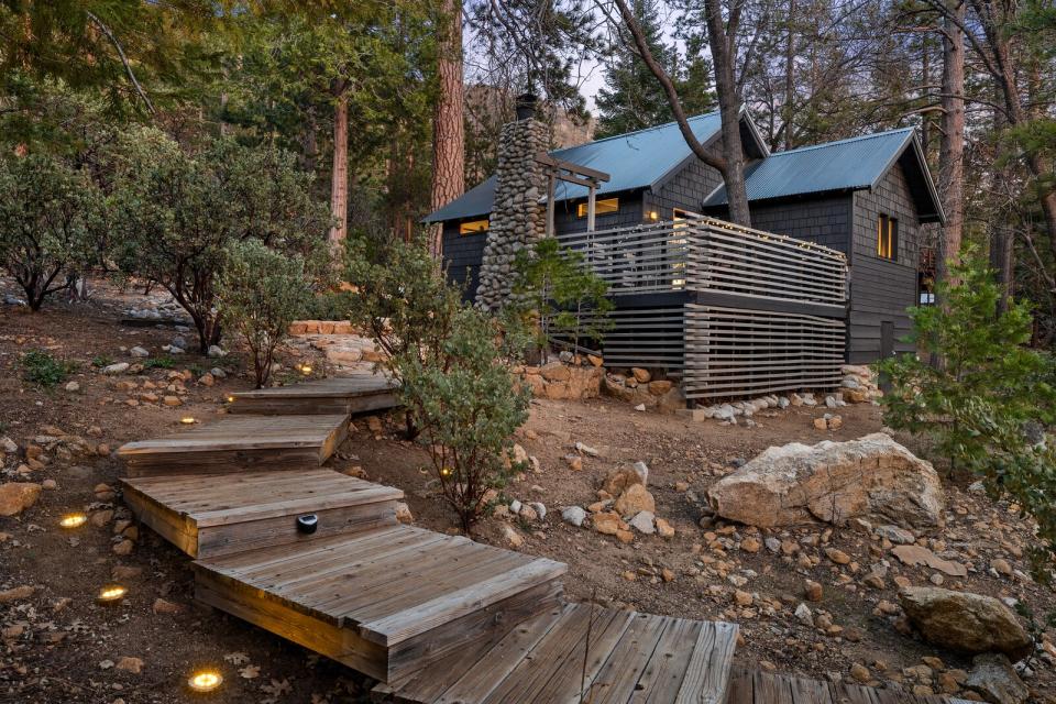 Wide wooden steps lead up to the a large outdoor deck, which has been carefully built around a soaring tree. The stone chimney pops against the home's sleek black exterior.