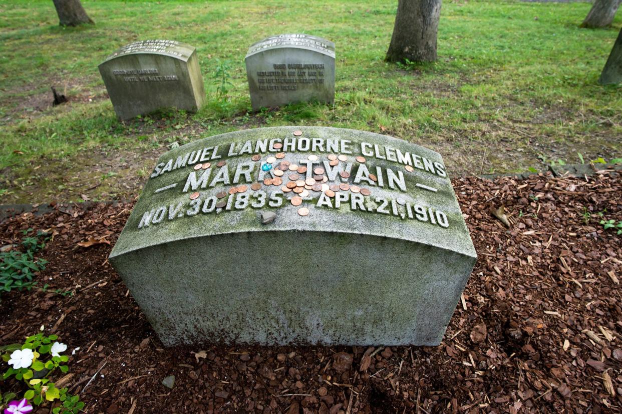 Mark Twain's grave in the Woodlawn Cemetery, Elmira, New York, with several coins on top on the words of the headstone with two headstones, a few trees and grass in the background