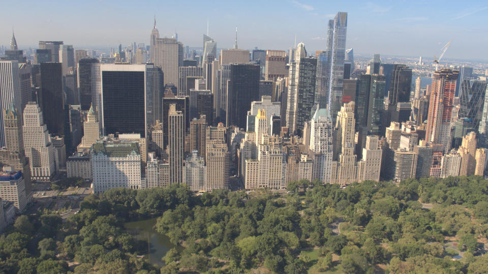 AERIAL ESTABLISHING SHOT: Flying above Central Park along 5th Avenue and towards Downtown Manhattan in sunny New York City. Tall glassy skyscrapers and condominium buildings overlooking Central Park