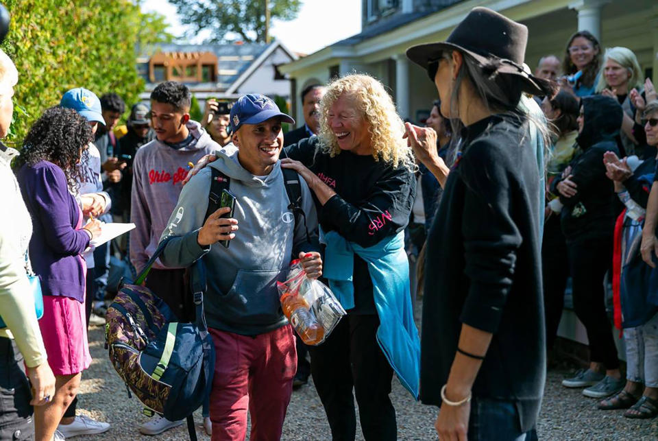 As migrants gather outside the church, with a line of residents looking on, a Venezuelan migrant wearing a backpack smiles shyly as an older woman puts her arm around his shoulder and another talks to him.
