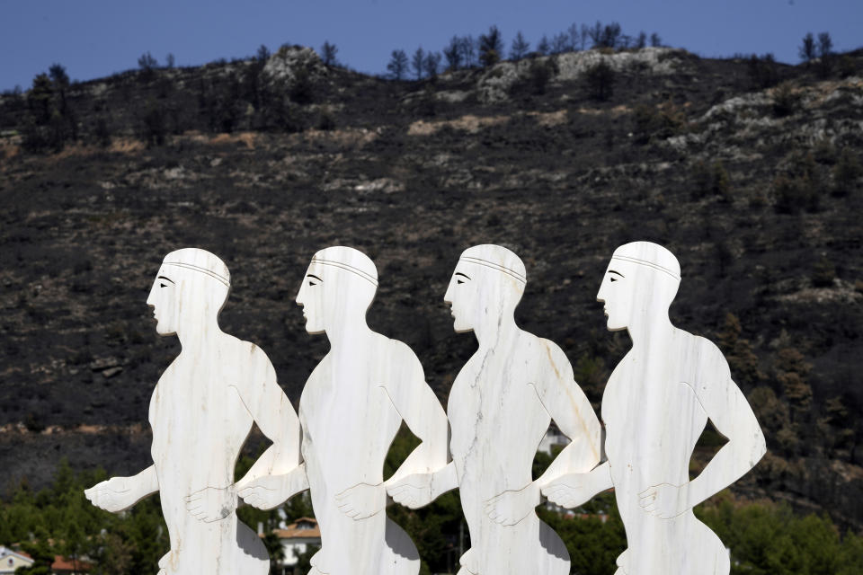 A sculpture, depicting marathon runners, stands in front of a blackened hill which had been burned in a mid-August wildfire, at Dionysos northern suburb of Athens, Thursday, Aug. 22, 2024. (AP Photo/Thanassis Stavrakis)