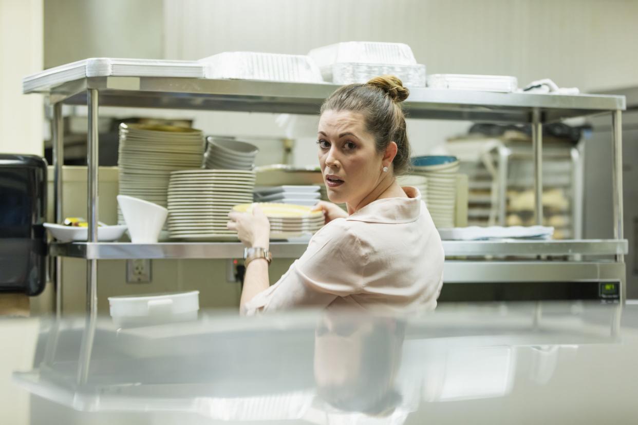 A mid adult Hispanic woman in her 30s working in a restaurant. She is in the kitchen, pulling a stack of plates from a shelf.