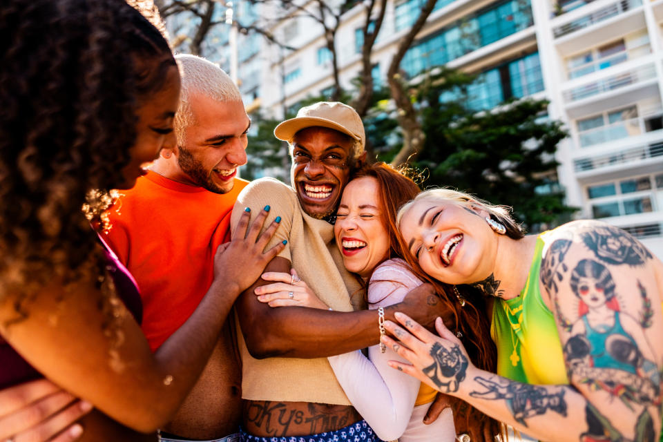 Group of friends, including noticeable LGTBQ+ representation, smiling and hugging outdoors, creating a sense of joy and inclusivity. Names not provided