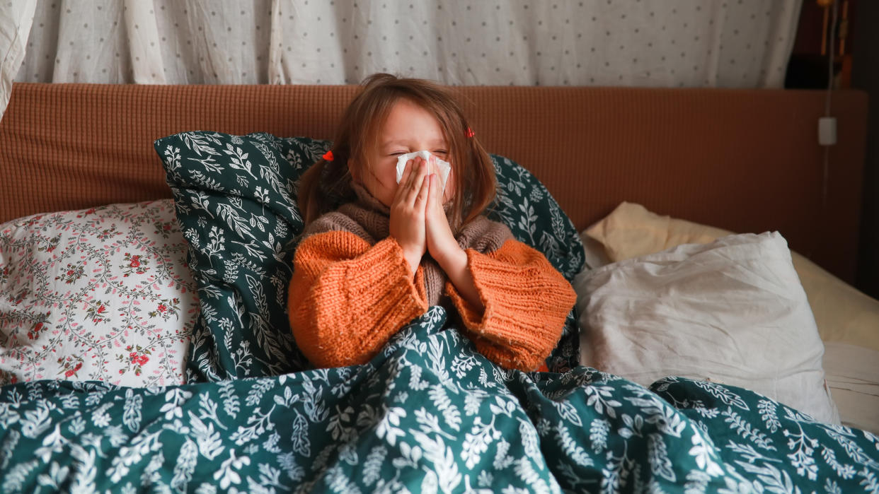 Sick Child Girl in Sweater Resting in Bed with a Tissue. Child Blowing Nose Covered in Blankets. Cold Season. Little Girl Cleaning Her Nose