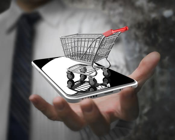 A man holding a smartphone with a shopping cart on top of it.